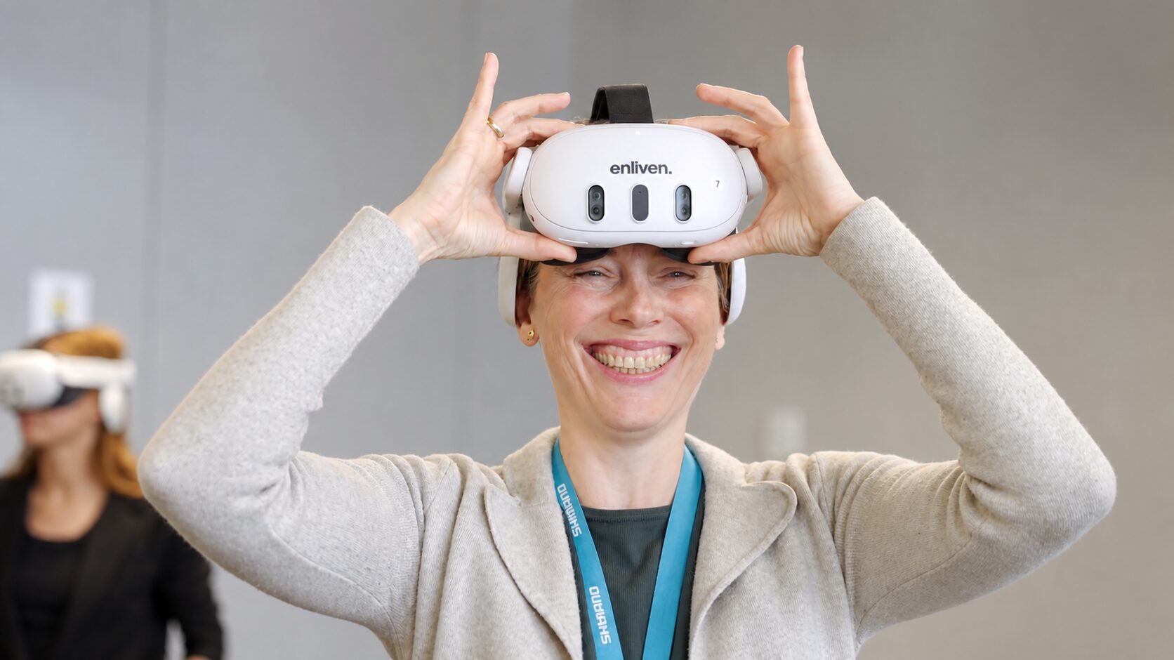 A smiling participant lifts an Enliven VR headset after an immersion, with another participant in a headset in the background.